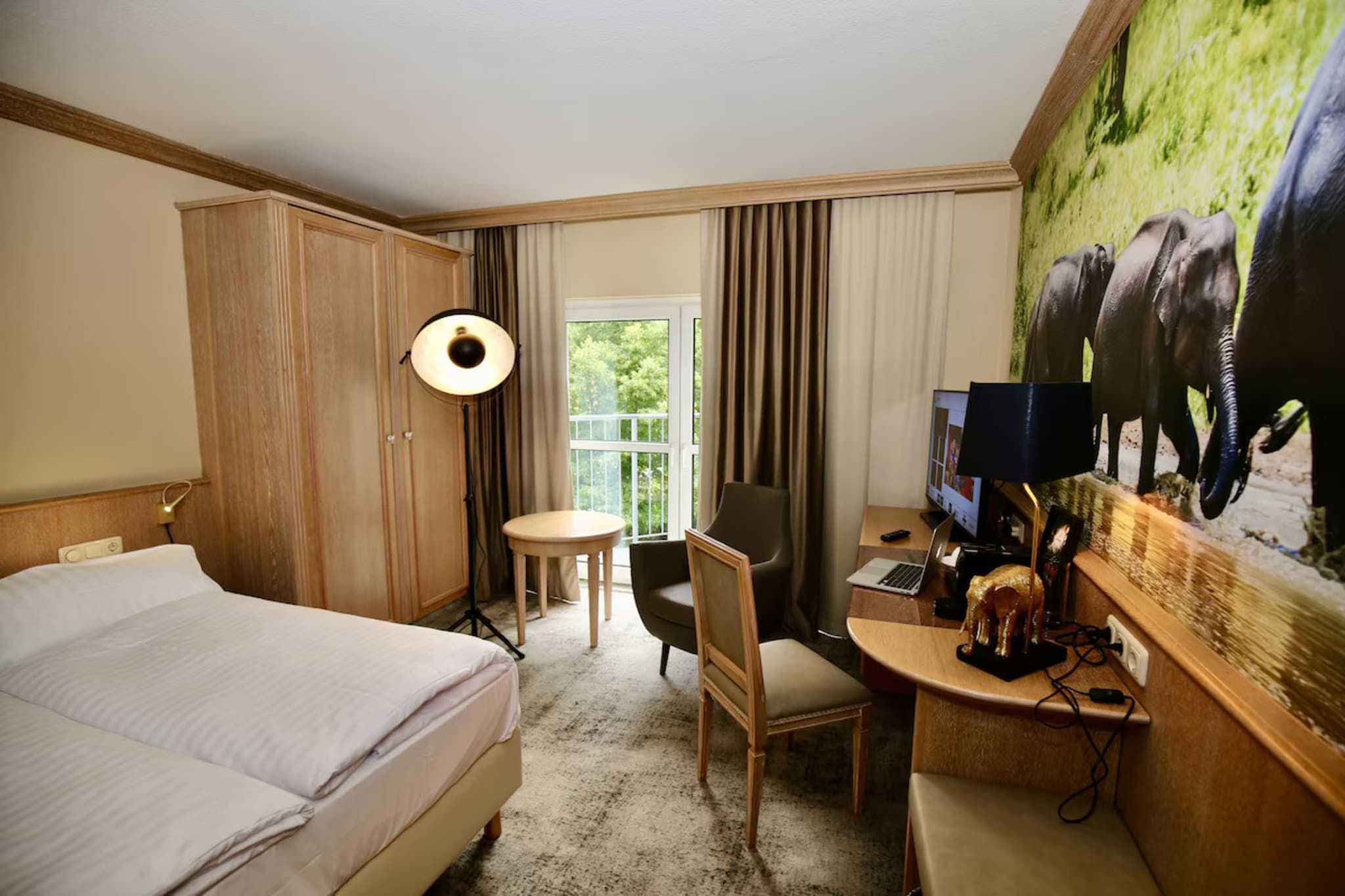 Ein Hotelzimmer mit einem Bett, einem Schreibtisch mit Computer, einem Stuhl, einer Lampe, einem Fernseher und einem großen Elefantenbild an der Wand.