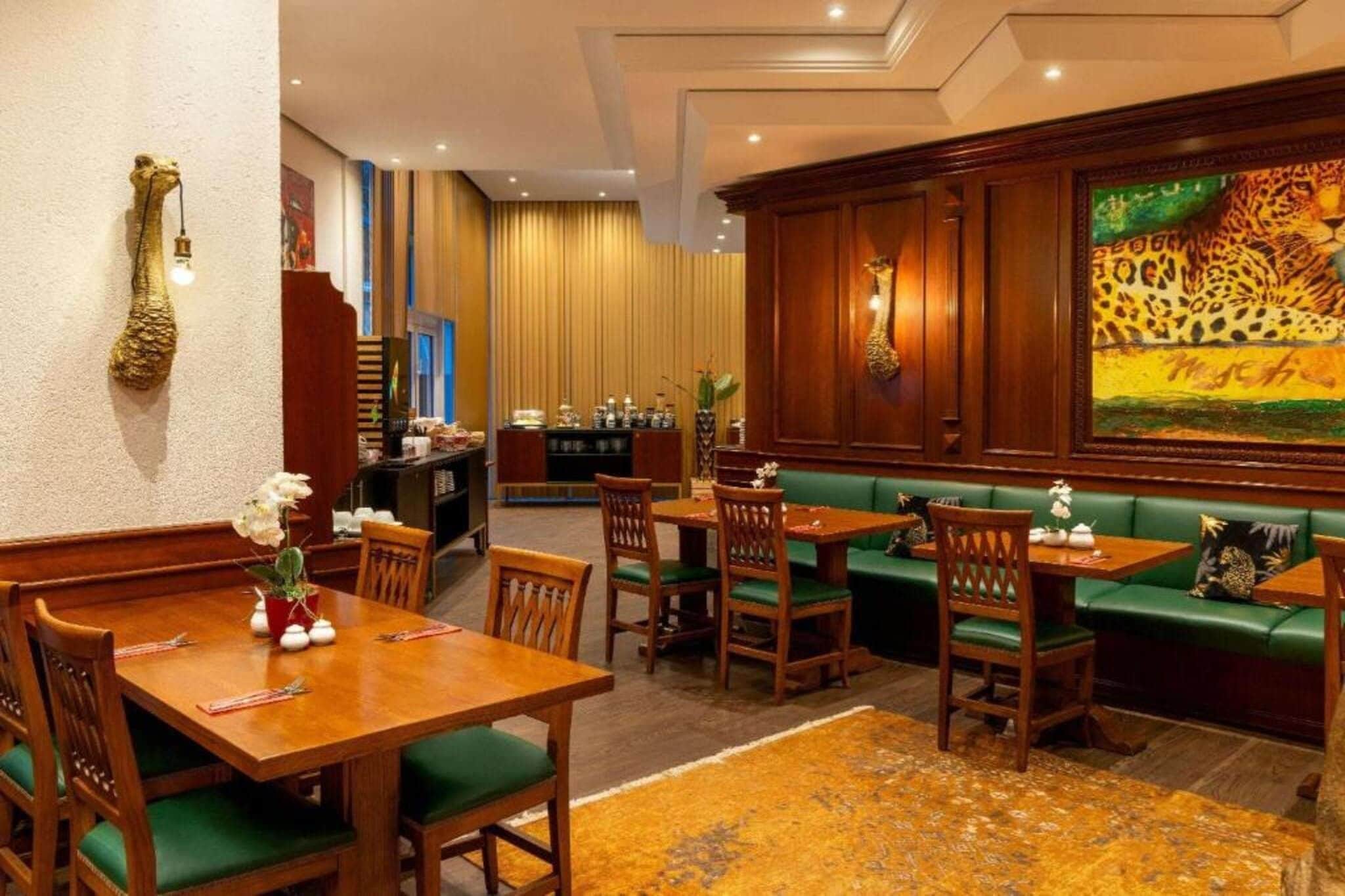 Een restaurantgedeelte in een hotel met houten tafels en zitplaatsen met groene kussens, een bar met een luipaard schilderij, en een decoratief wandobject.