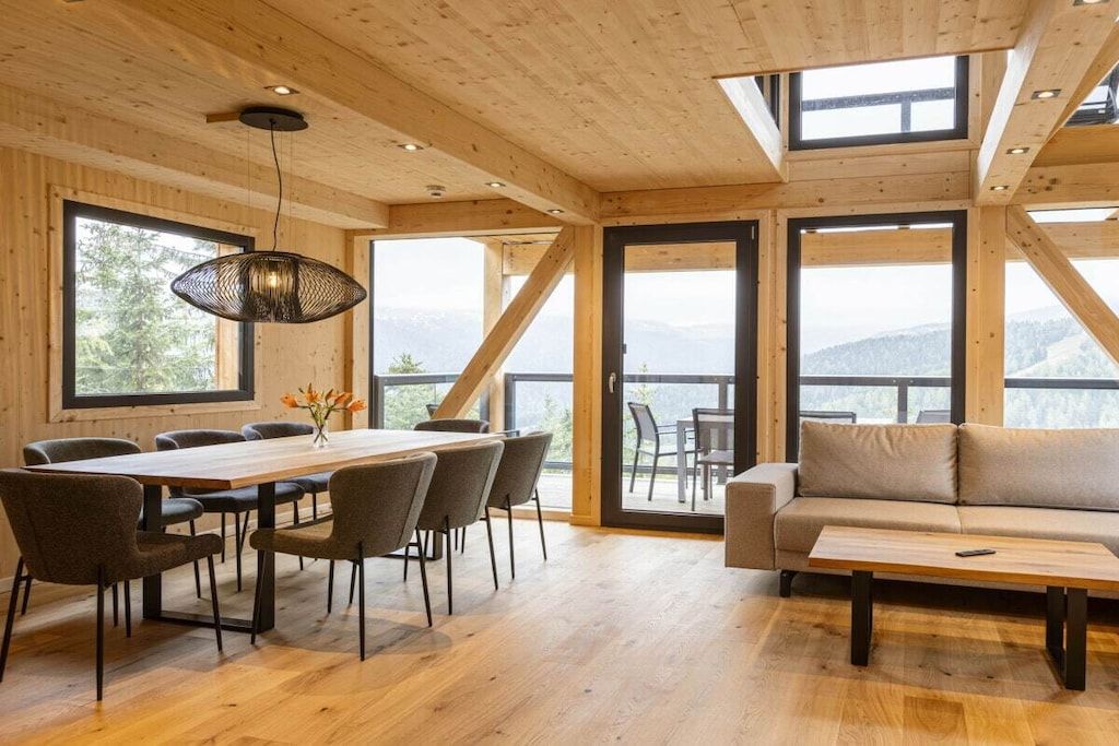 Un intérieur en bois comprenant une table à manger, des chaises, un canapé et de grandes fenêtres donnant sur l’extérieur.