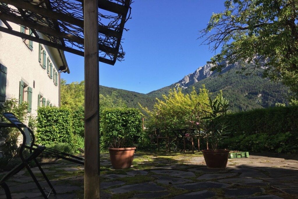 Un patio con piso de piedra, plantas en macetas, una silla de metal y un edificio con una pérgola de madera.