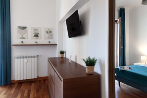 Habitación de hotel con cómoda de madera, televisor de pantalla plana montado en la pared, cama con colcha color turquesa y ventana con cortinas azules.