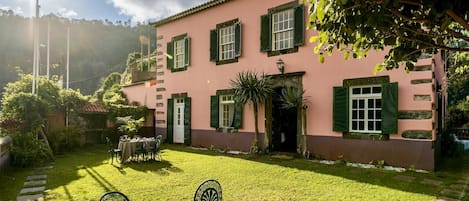 A pink building with green shutters, a patio with a table and chairs, and a garden with a pathway.