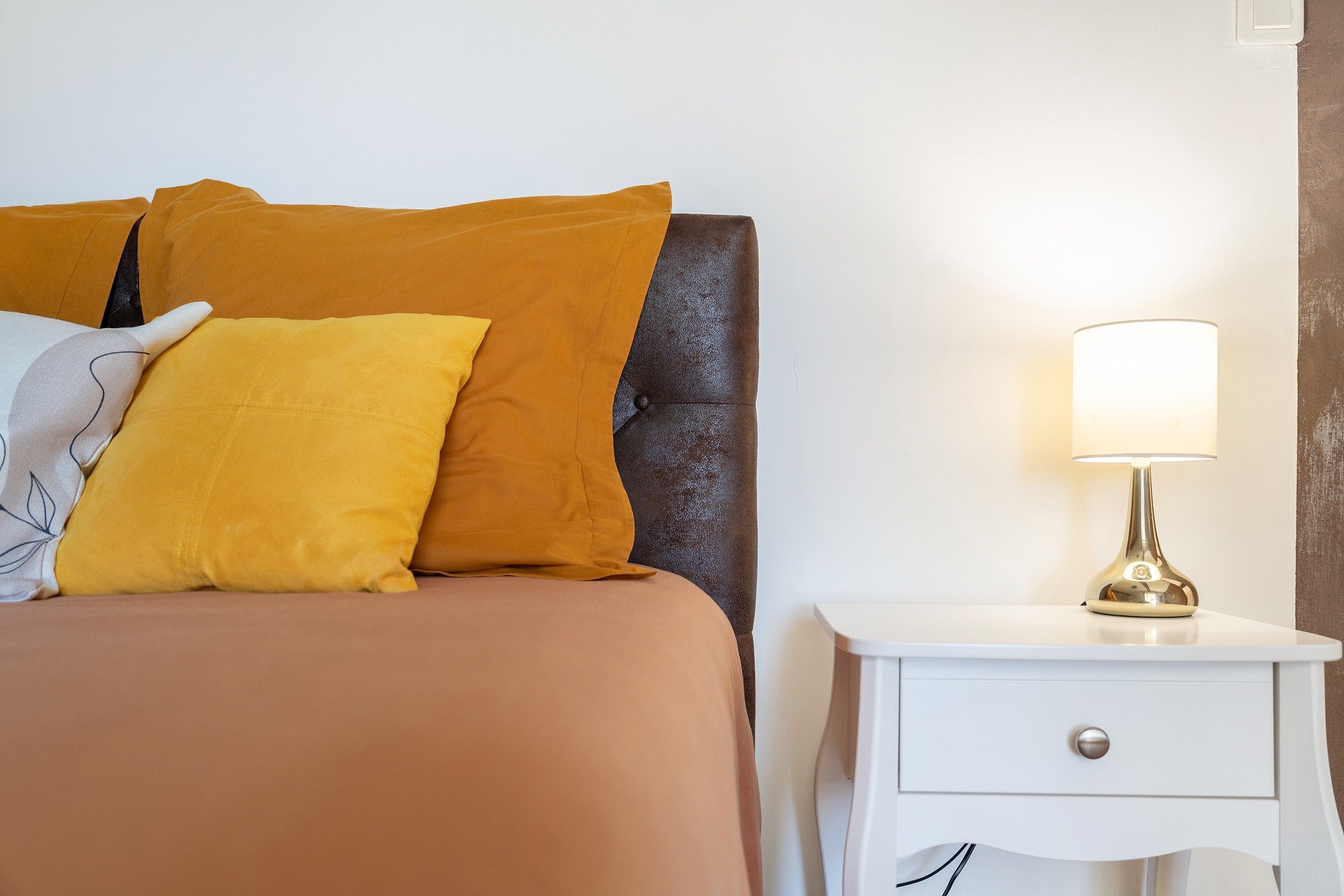 Un lit avec une tête de lit marron et plusieurs oreillers, une table de chevet avec une lampe et une commode blanche.