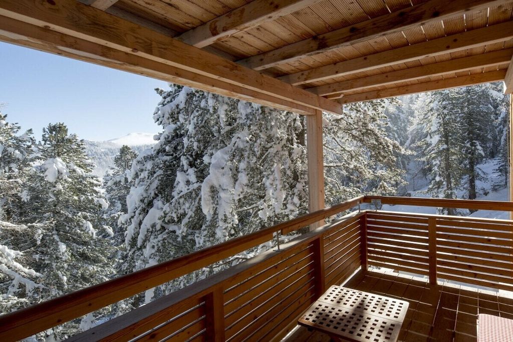 A wooden balcony with a metal bench, overlooking a snowy forest.
