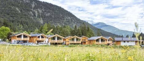A row of wooden cabins with balconies, set in a grassy field with mountains in the background.