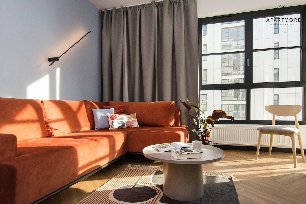 Ein modernes Wohnzimmer mit einer Eckcouch, einem Couchtisch, einem Sessel und einem großen Fenster mit Blick auf Gebäude.