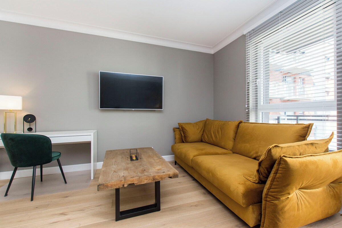 Ein Wohnzimmer mit flachbildfernseher an der Wand, einem Holztisch, einem senfgelben Sofa, einem grünen Sessel und einem weißen Schreibtisch.