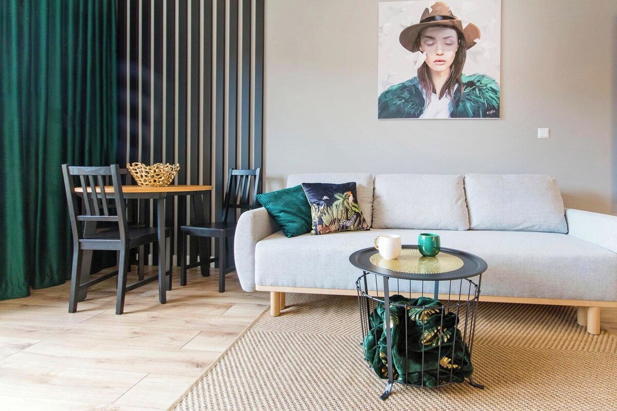 Ein modernes Wohnzimmer mit einer Couch, zwei Sesseln, einem kleinen Tisch mit Tasse und grüner Kanne, einem dekorativen Korb und einem gerahmten Bild an der Wand.
