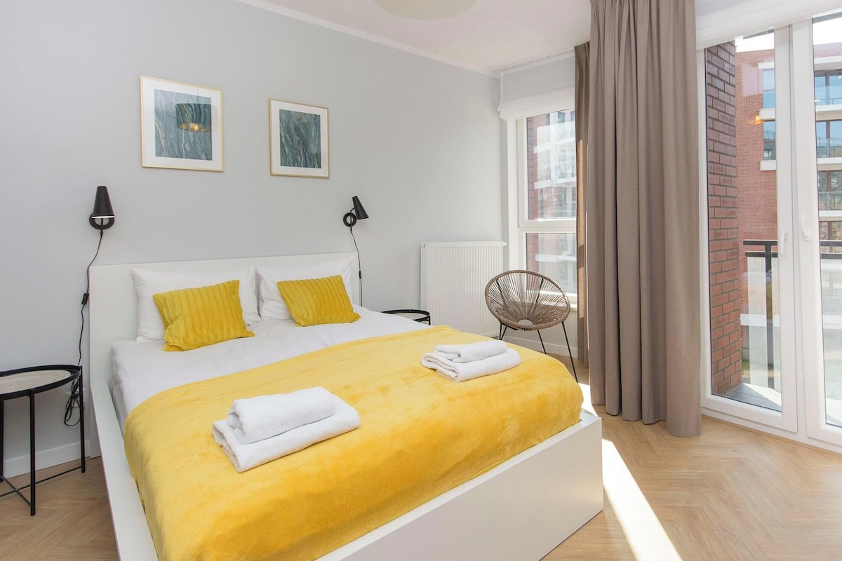 Ein modernes Schlafzimmer mit einem großen Bett, zwei Nachttischlampen, einem Sessel und Blick auf die Stadt durch große Fenster.