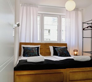 A double bed with a wooden headboard, white bedding, and two decorative pillows. There is a window with white curtains, a bedside table with a lamp, and a clothing rack to the right. - Shipyard Apartments by Apartmore (Gdańsk)