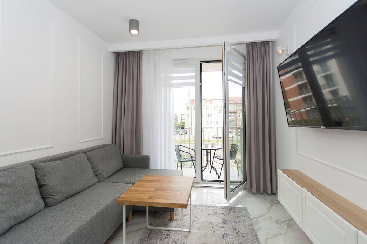 Ein modernes Wohnzimmer mit einem grauen Sofa, einem Holztisch, einem an der Wand montierten Fernseher und einem Balkon mit Blick auf Gebäude.