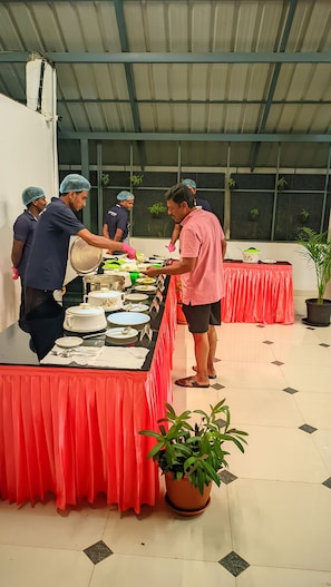 A buffet setup with multiple serving stations, a tiled floor, and a potted plant. - Grace Grand Resort Munnar (Devikolam)