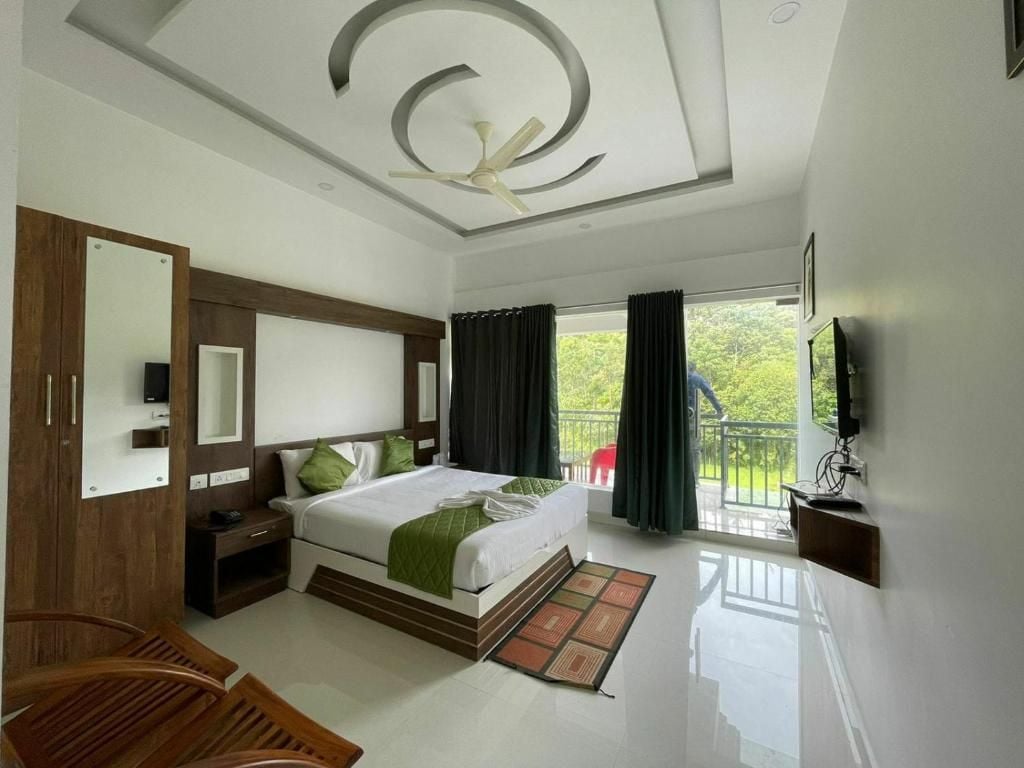 A hotel room with a bed, a TV, a ceiling fan, a balcony, and a view of greenery.
