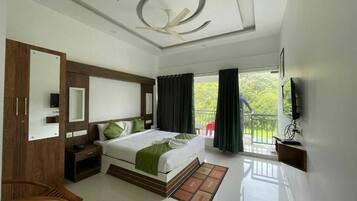 A hotel room with a bed, a TV, a ceiling fan, a balcony, and a view of greenery.