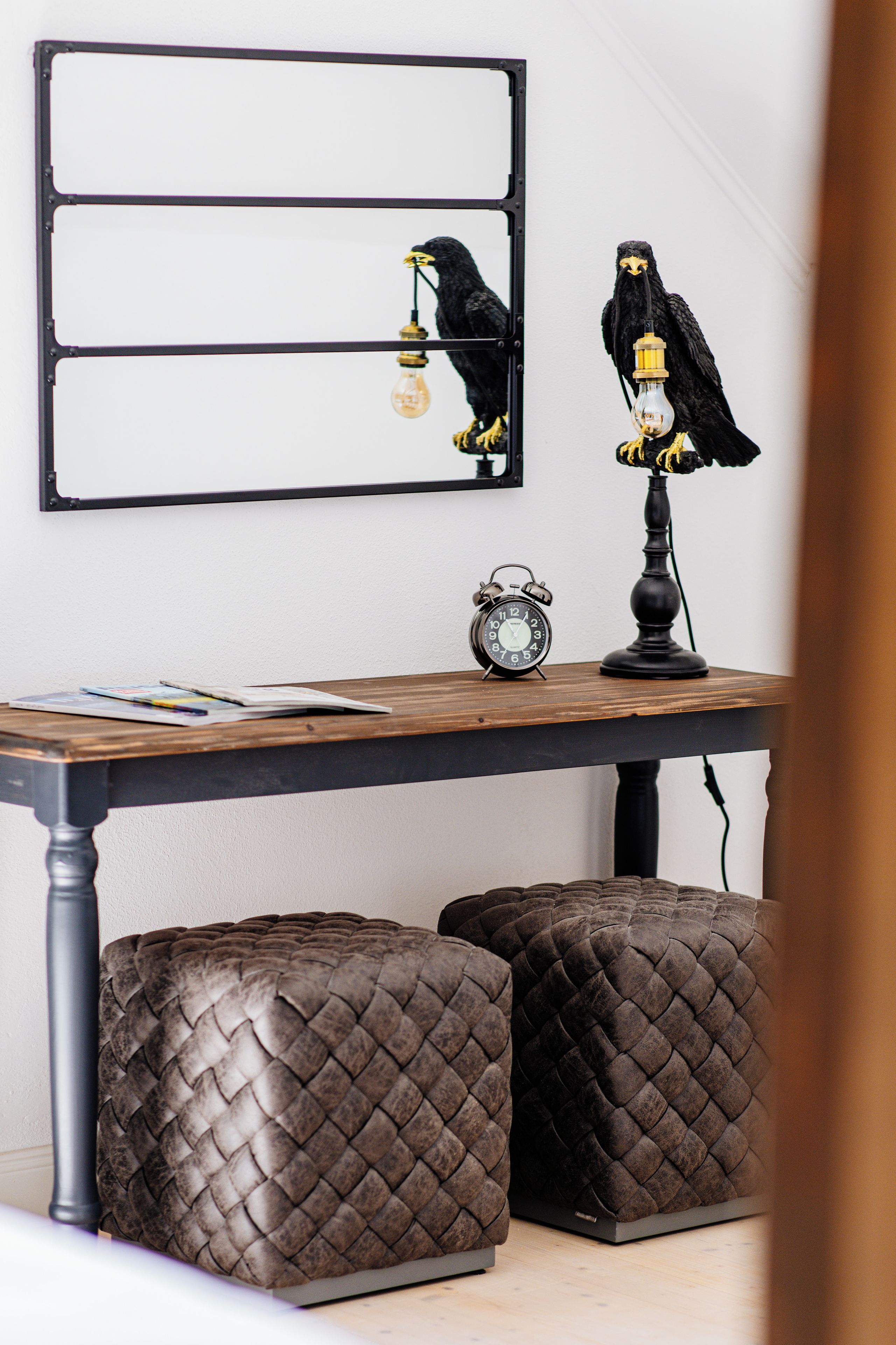 Une table à manger avec un cadre en métal noir, deux poufs noirs à texture tissée, une horloge murale noire et une figurine d’oiseau noir tenant une ampoule.