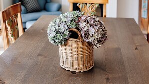 A dining table with a wicker basket containing flowers, a blue sofa, a fireplace, and a chandelier.