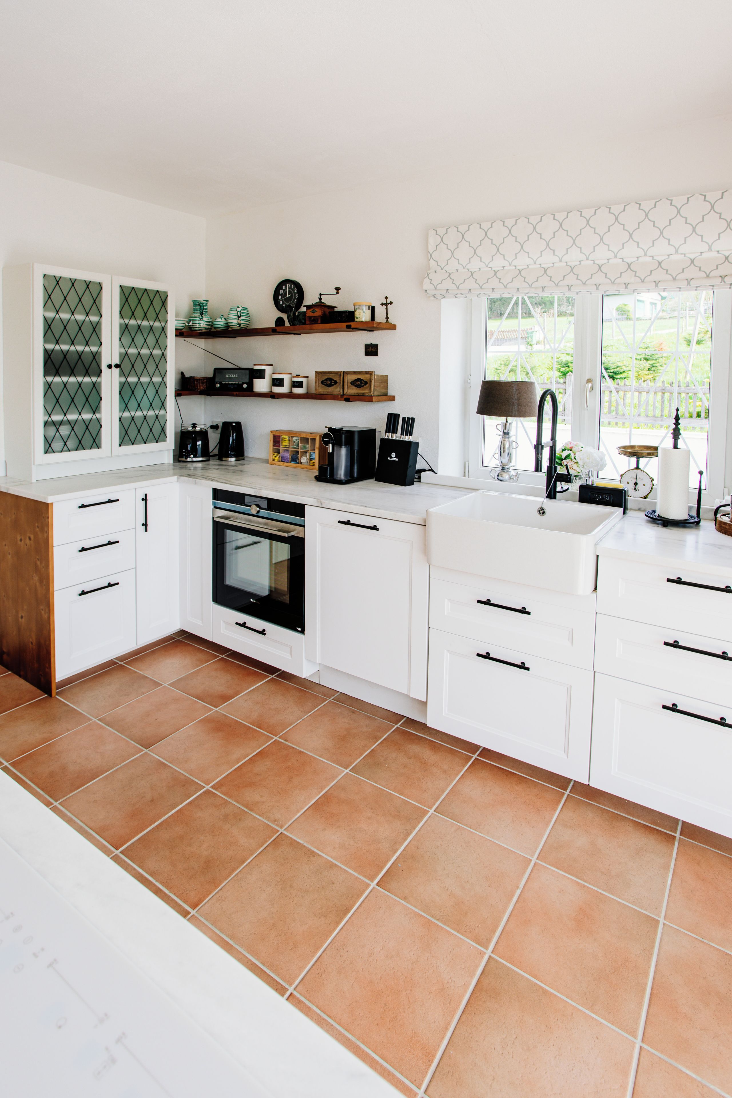 Une cuisine moderne avec des meubles blancs, un four encastré et une grande fenêtre donnant sur un paysage verdoyant.