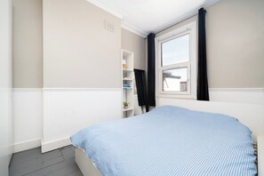 Ein Schlafzimmer mit Bett, einem Fenster mit Vorhängen, einer weißen Regalwand und einer kleinen Pflanze.