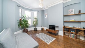A living room with a sofa, a television, wooden flooring, a chandelier, and a bookshelf. - Bright Garden Flat with Private Outdoor Space (Greater London)