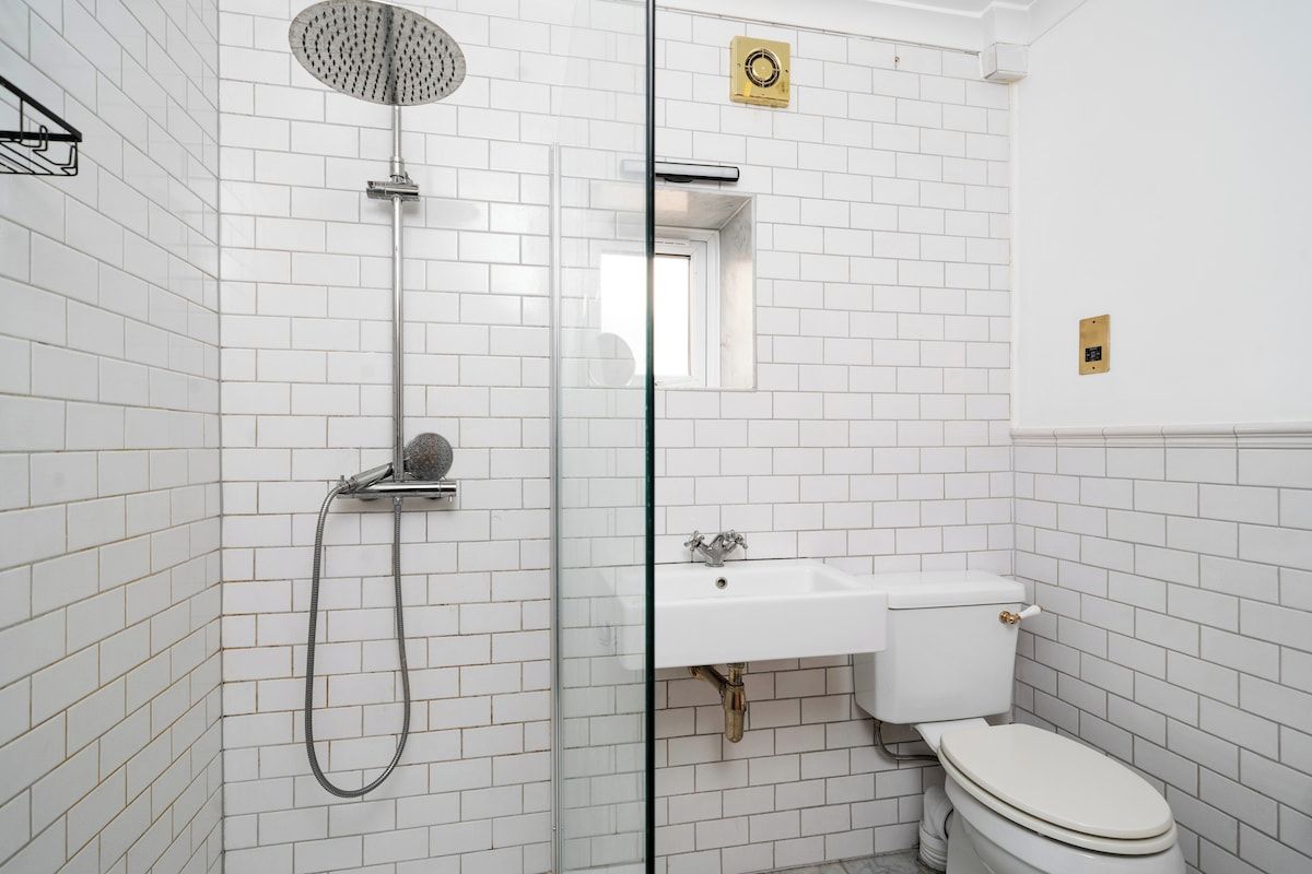 Bany amb parets de rajola blanca, dutxa amb envàs de vidre, un lavabo blanc i un vàter.