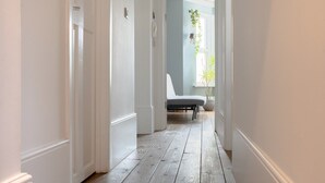 A hallway with white walls and wooden flooring leading to a room with a bed and a hanging plant. - Bright Garden Flat with Private Outdoor Space (Greater London)