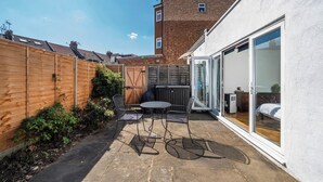 A patio area with a table and chairs, a wooden fence, and a building with a brick facade in the background. - Bright Garden Flat with Private Outdoor Space (Greater London)