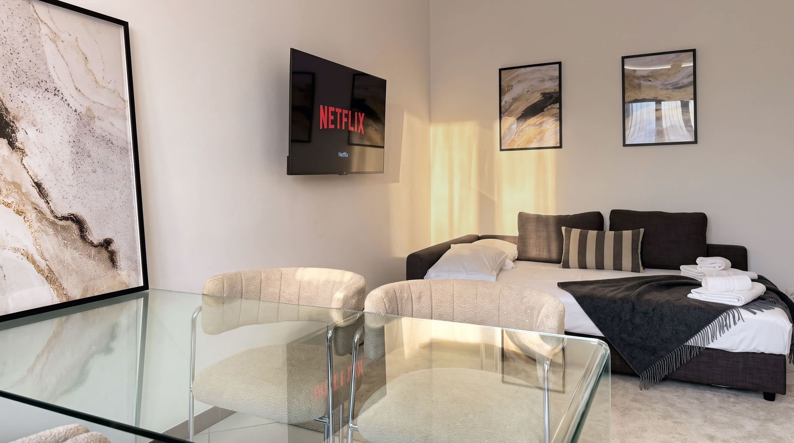 A modern hotel room with a glass table, a bed with a dark headboard, and artwork on the walls.