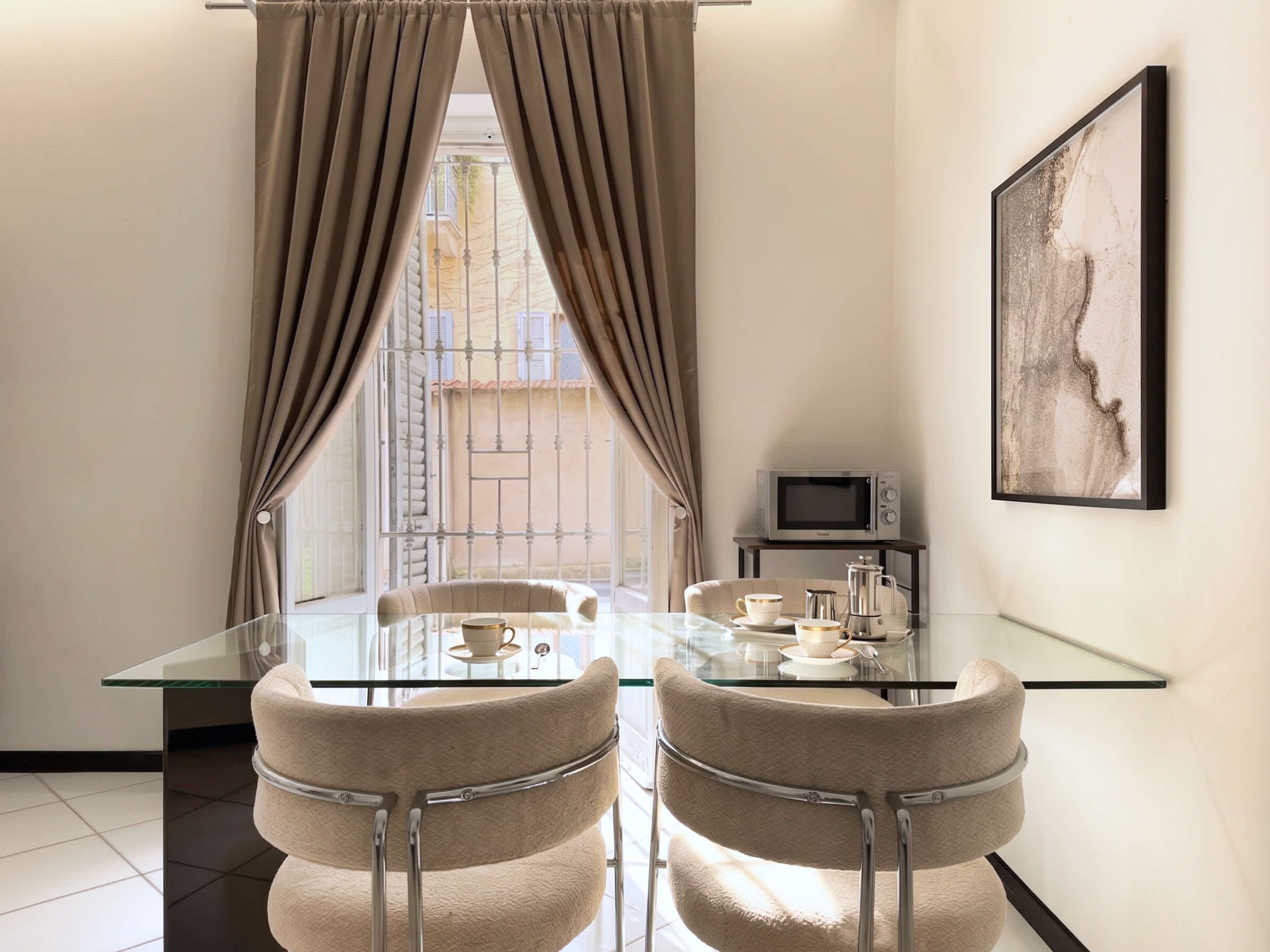 Un comedor moderno con una mesa de vidrio, dos sillas acolchadas, una pequeña kitchenette con microondas y una obra de arte enmarcada en la pared.