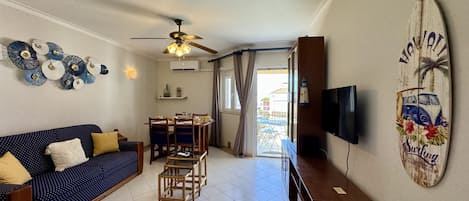 A living room with a sofa, a dining table, chairs, a ceiling fan, a TV, and a surfboard decoration.