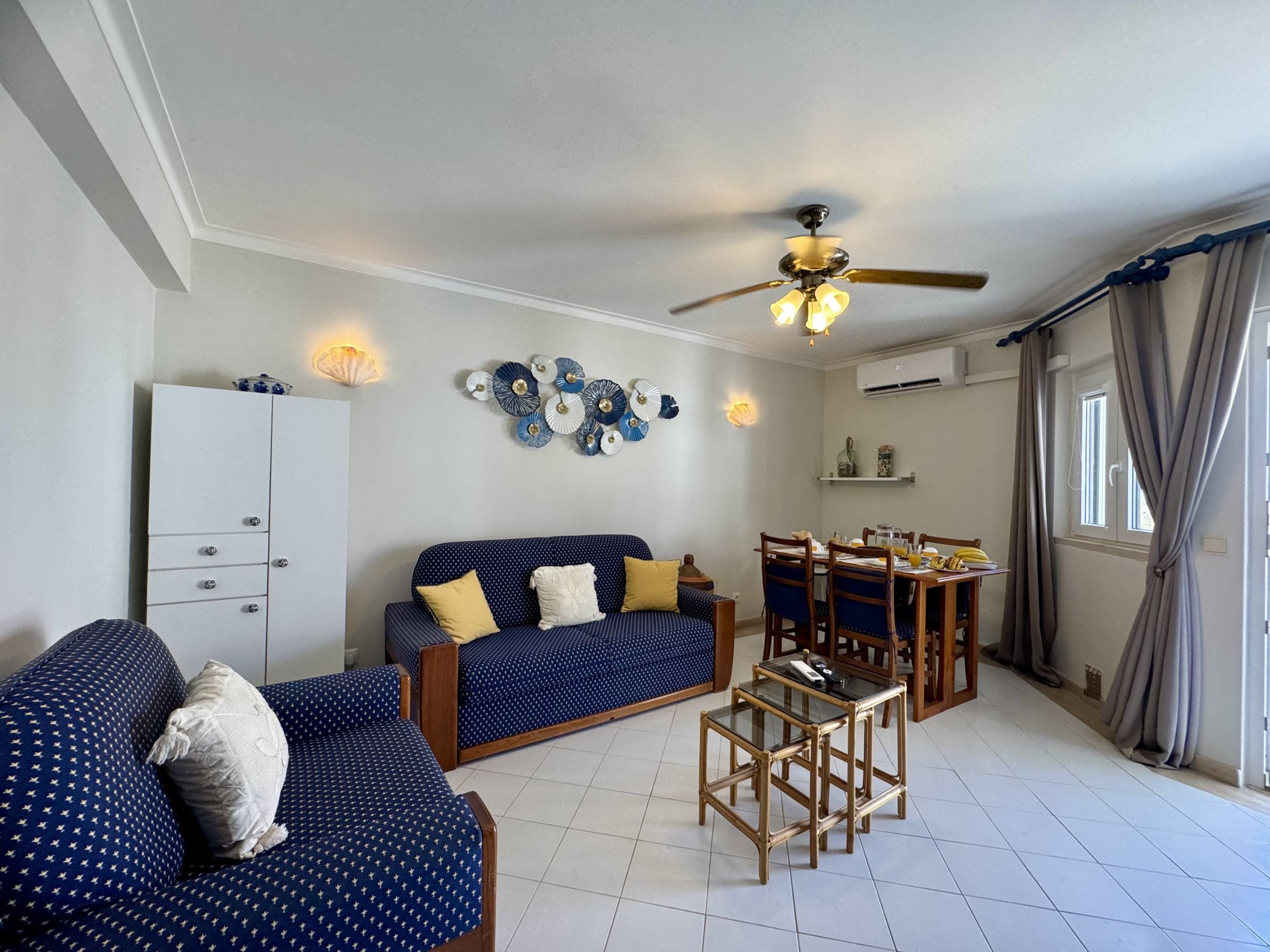 A living room with a sofa, a dining table with chairs, a ceiling fan, decorative wall art, and a cabinet.