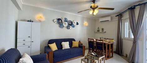 A living room with a sofa, a dining table with chairs, a ceiling fan, decorative wall art, and a cabinet.
