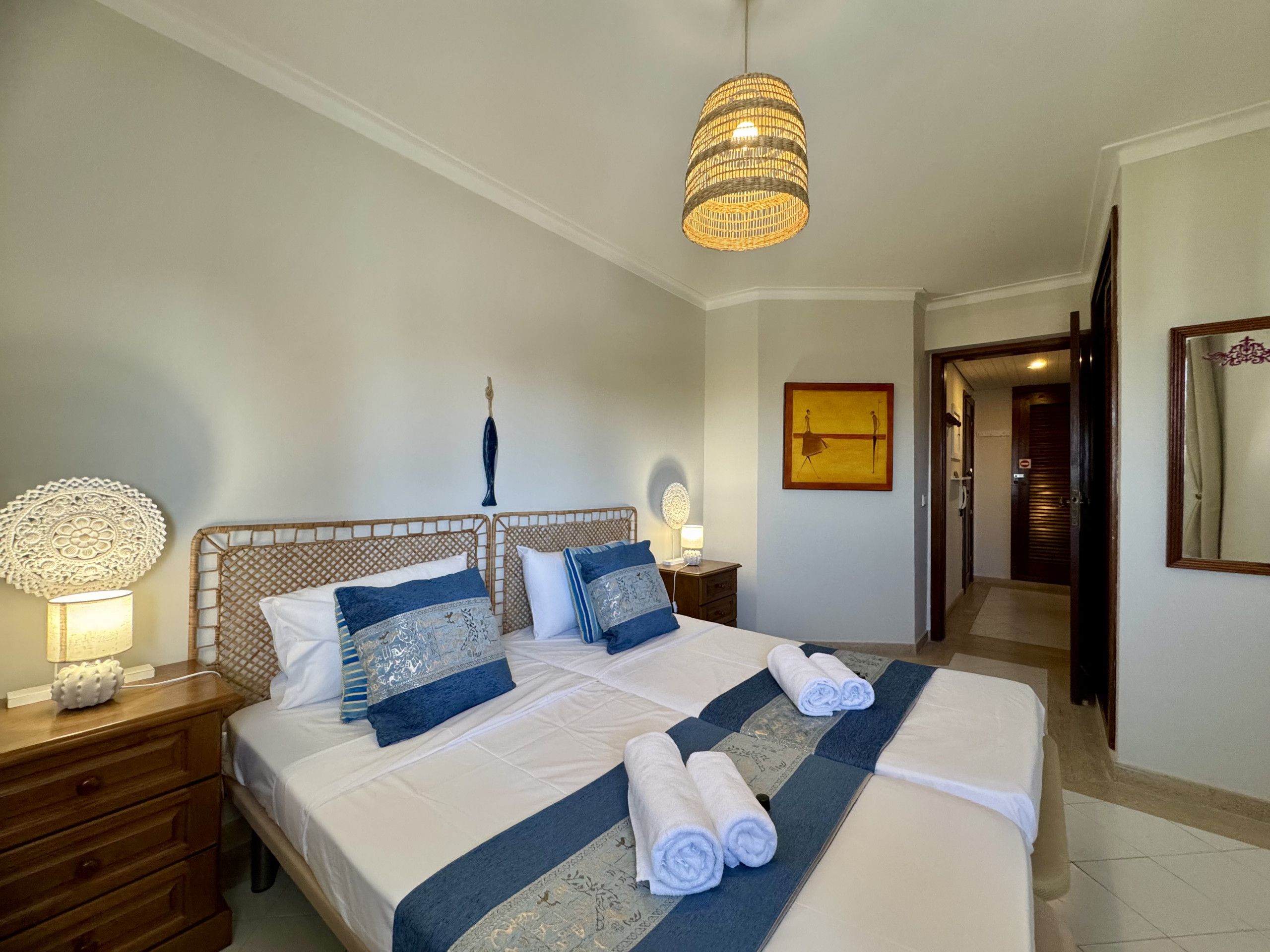 A hotel room with a large bed, a bedside table with a lamp, a wall-mounted artwork, and a door leading to another room.