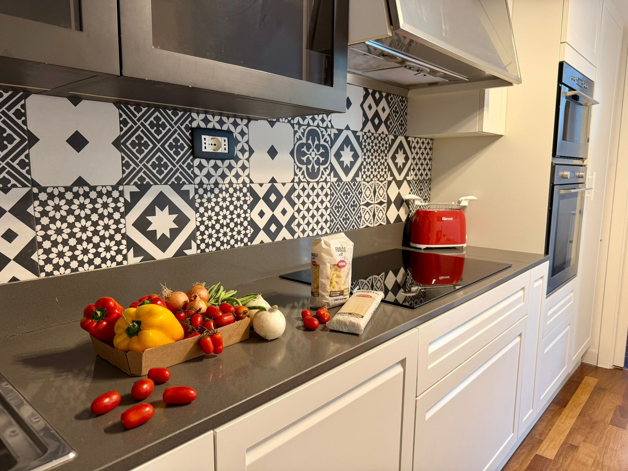 Et køkken med et mønstret backsplash, en rød toaster og diverse friske grøntsager på bordet.