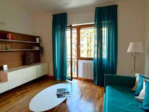 Una sala de estar con un sofá blanco, un sofá color turquesa, una mesa de centro y una lámpara. Se observa una estantería con libros y objetos decorativos.