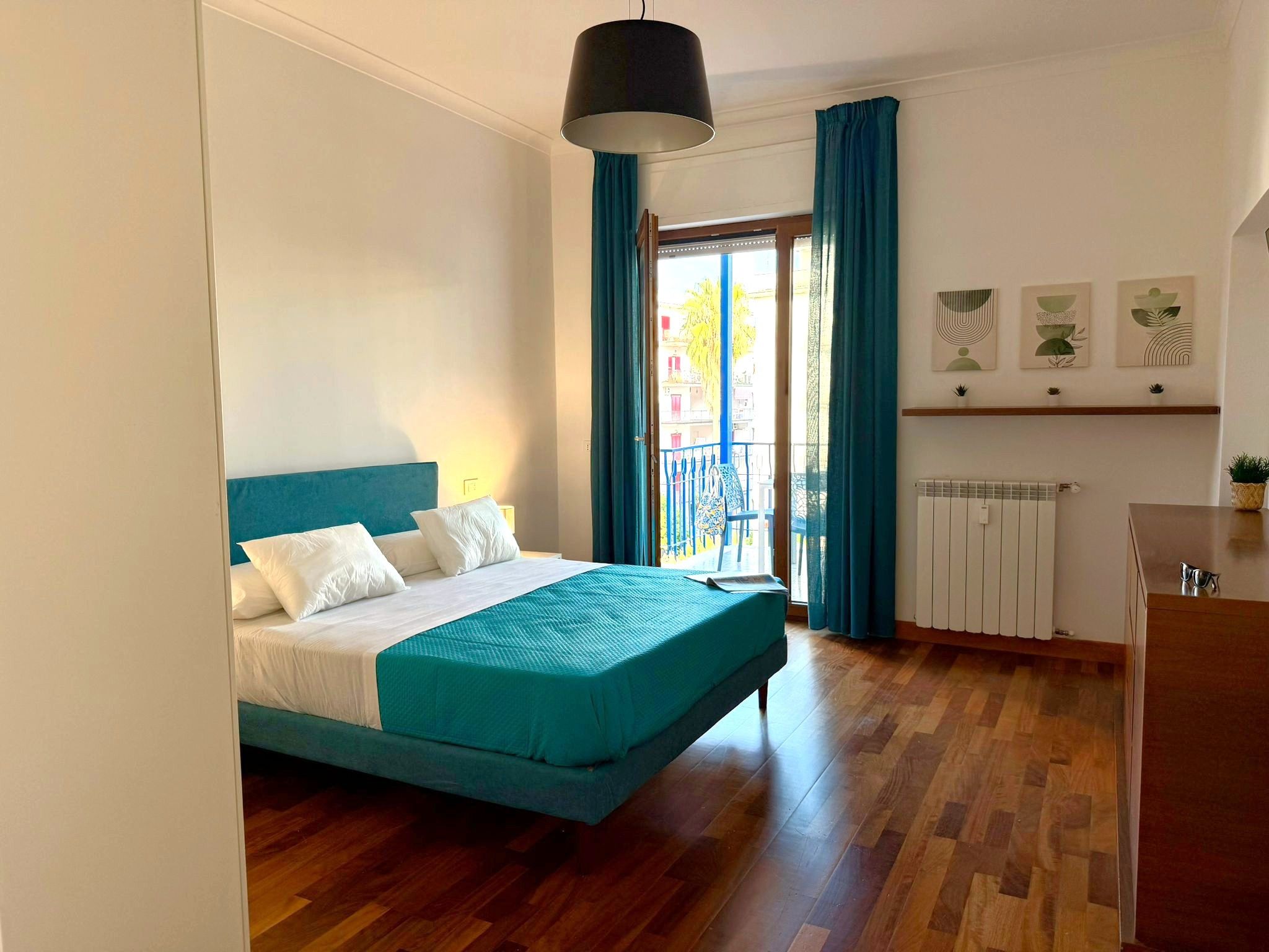 Een slaapkamer met een bed, een radiator, een raam met gordijnen en uitzicht op gebouwen.