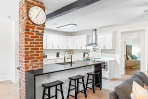 A modern kitchen with white cabinets, a brick accent wall, a central island with stools, and a visible clock. - Pet-friendly Pensacola Escape - Sleeps 10 (Pensacola)