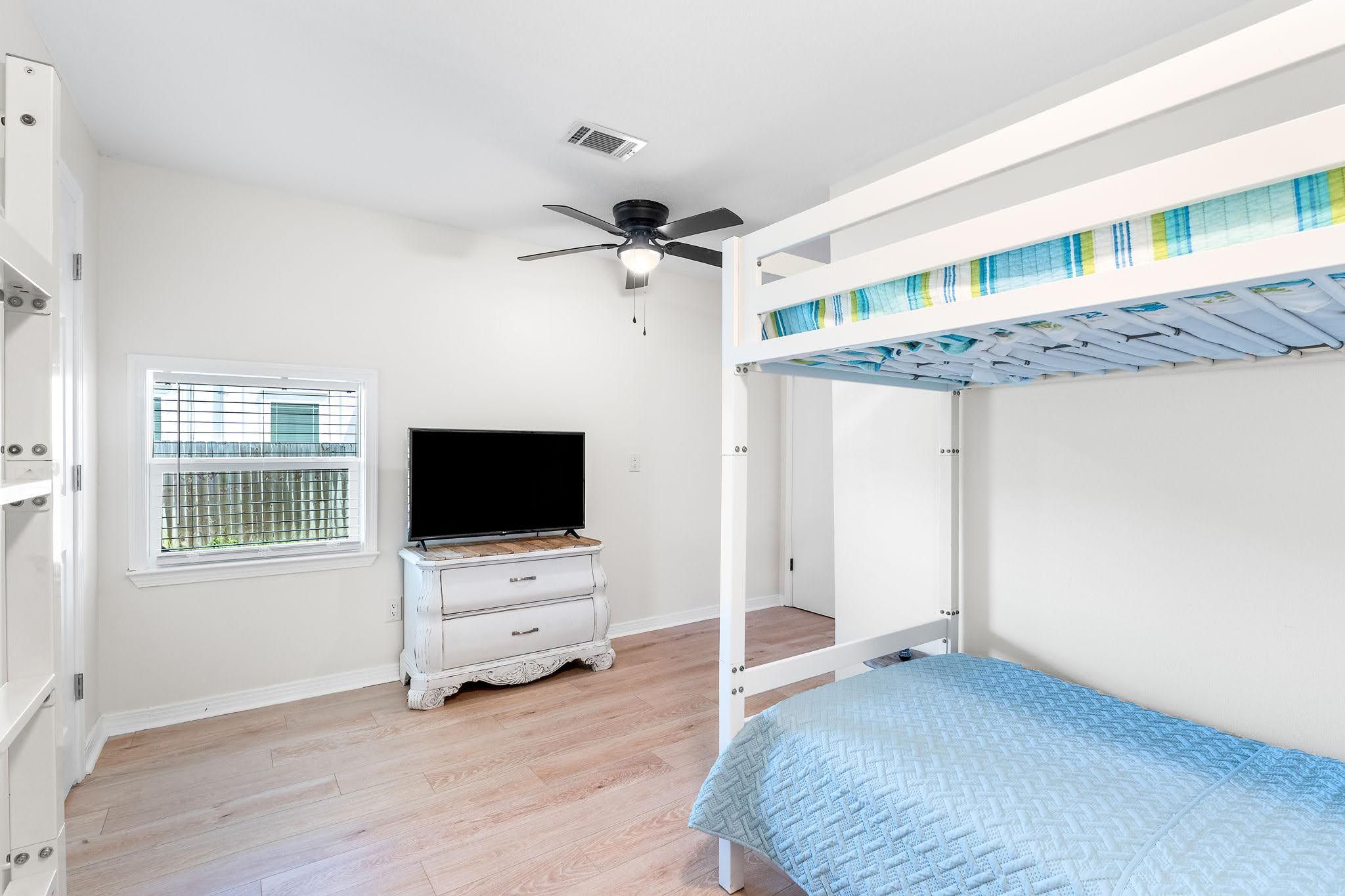 Ein Zimmer mit einem Etagenbett, einem Fernseher auf einer Kommode, einem Fenster mit Jalousien und einem Deckenventilator.