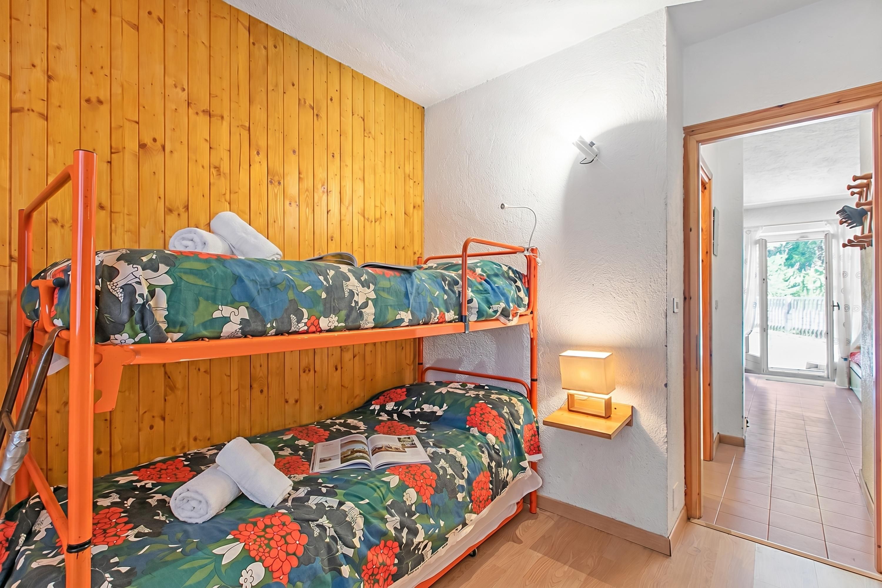 Un lit superposé avec une literie à motifs floraux, un mur en bois, une petite table de chevet avec une lampe et une porte donnant sur un espace extérieur.