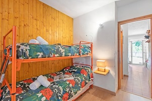 Uma cama beliche com roupa de cama florida, uma parede de madeira, uma mesinha de cabeceira com abajur e uma porta que dá para uma área externa.
