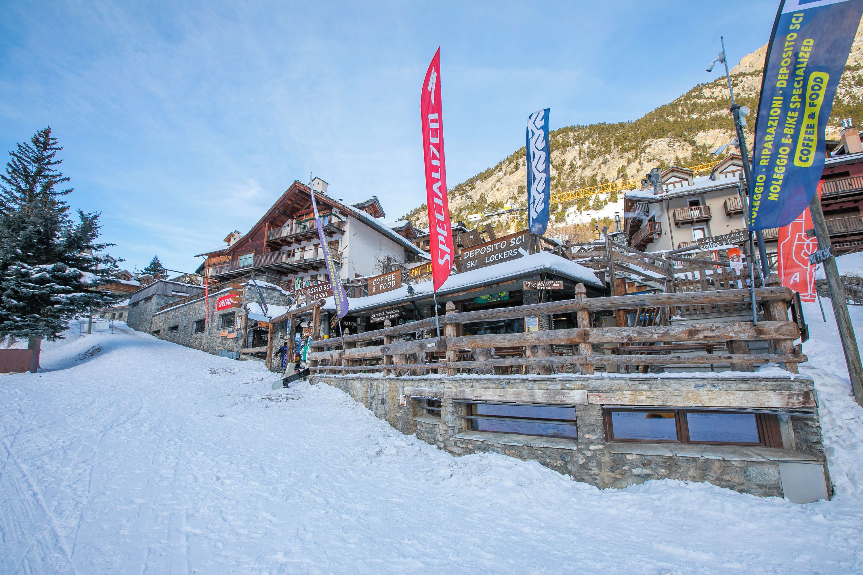 Un pueblo en la montaña cubierto de nieve, con edificios de madera y banderas que señalan servicios como taquillas para esquíes y cafetería.