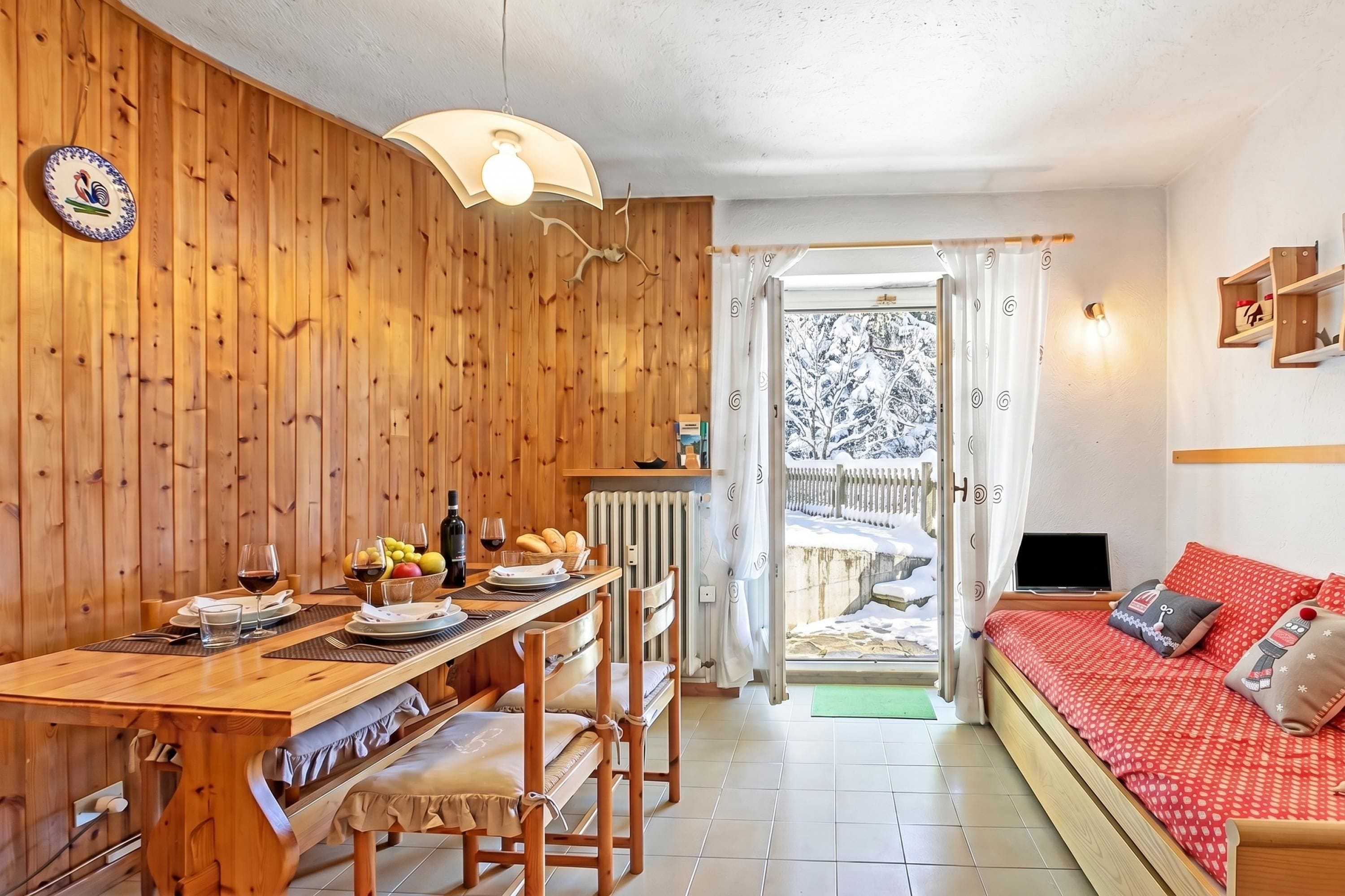 A wooden interior with a dining area, a bed with red bedding, and a view of a snowy landscape through a glass door.