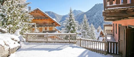A snow-covered wooden chalet with a balcony, surrounded by snow-laden trees and mountains in the background.