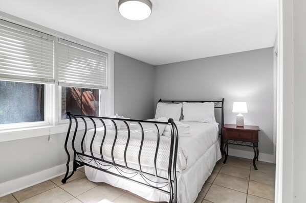 A bedroom with a bed, a bedside table with a lamp, and a window with blinds.