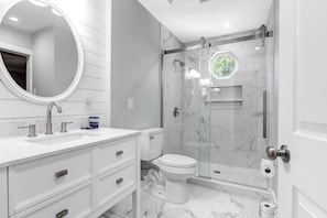 Un baño moderno con tocador blanco, un espejo redondo, inodoro y zona de ducha con mampara de vidrio.
