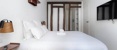 Una cama bien hecha con ropa de cama blanca, una mesita de noche de madera con un control remoto, un estante de pared con una planta y un televisor en la pared.