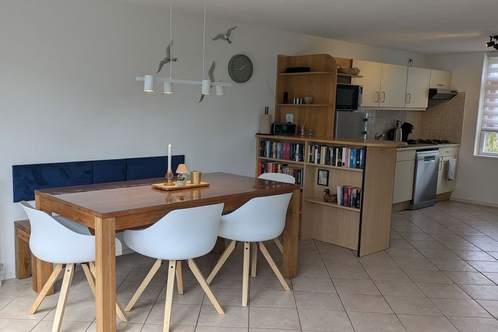 A modern kitchen with a dining area, featuring a wooden table, white chairs, a bookshelf, and a clock on the wall.