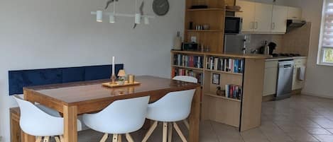 A modern kitchen with a dining area, featuring a wooden table, white chairs, a bookshelf, and a clock on the wall.
