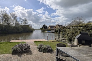 A riverside view with a wooden dock, a grassy area with shrubs, and a building with a sloped roof.