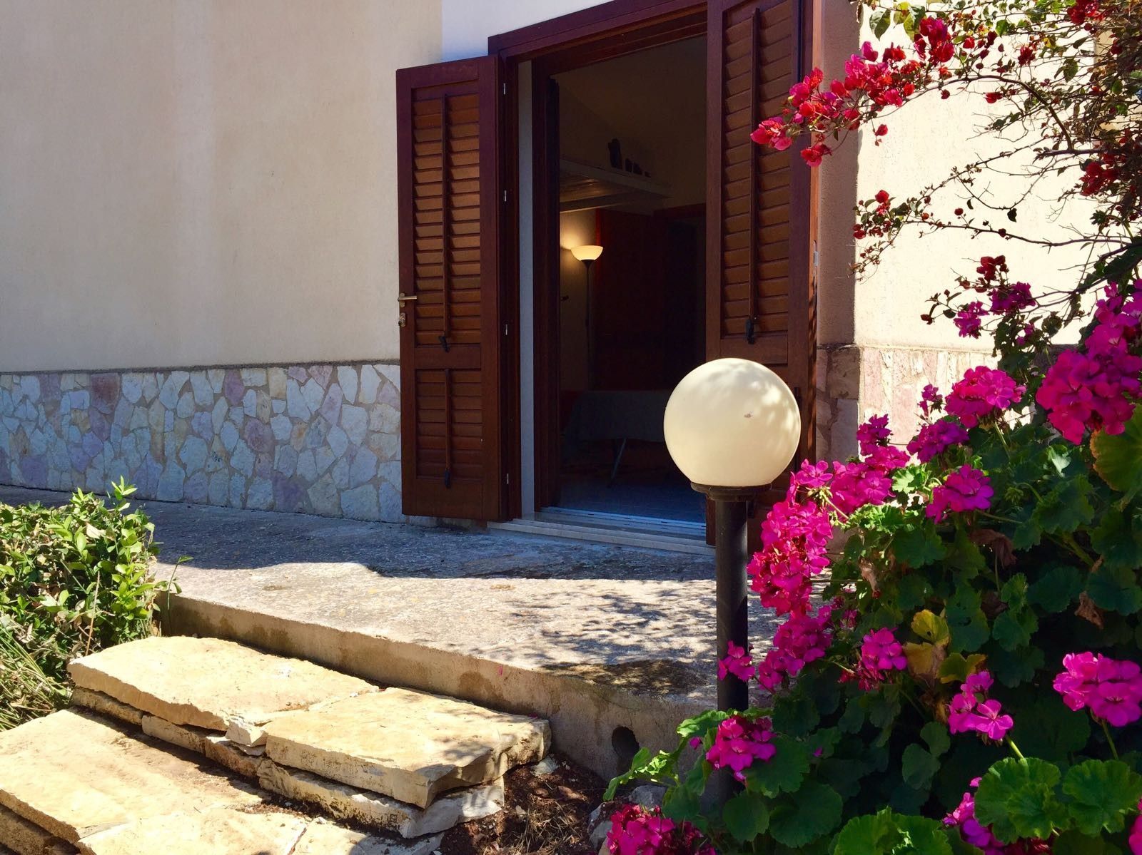 Una escalera de piedra que conduce a una puerta de madera con contraventanas, una lámpara y vibrantes flores rosas.