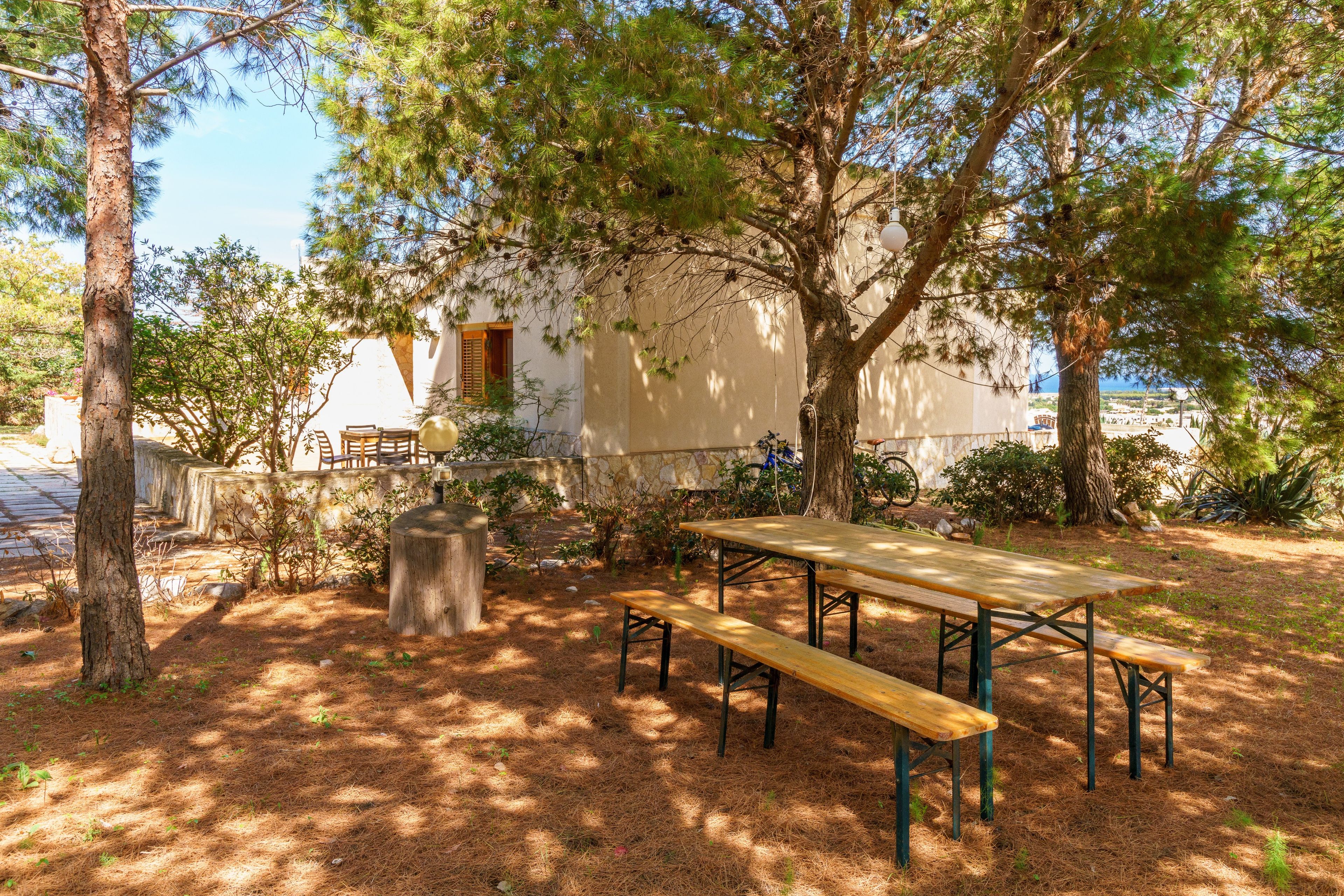 Un espace extérieur aménagé avec une table de pique-nique et des bancs, entouré d’arbres et bordé d’un bâtiment doté d’un mur en pierre.